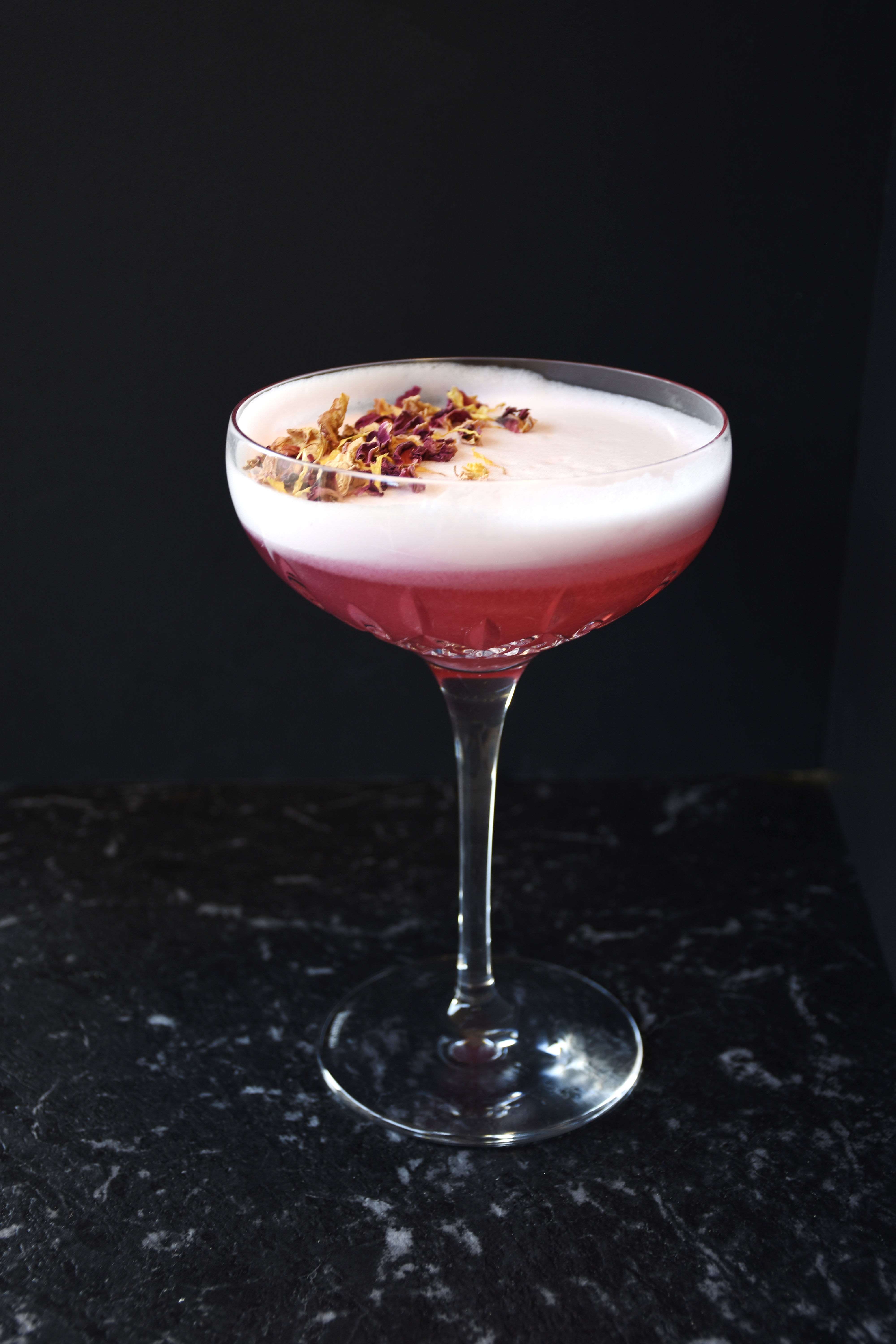 A pink cocktail with white foam and dried flower petals on top in a crystal cocktail glass against a black background